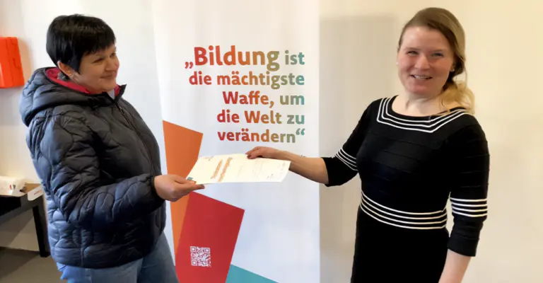 Dozentin Albina Müller übergibt einer Teilnehmerin am OWYO CAMPUS eine Teilnahmebescheinigung für einen abgeschlossenen Sprachkurs im Rahmen der AZAV-zertifizierten Bildungsangebote.